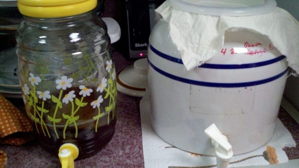 A glass jar with a yellow lid adorned with floral patterns, containing kombucha, next to a white ceramic crock with blue stripes, covered with a cloth.